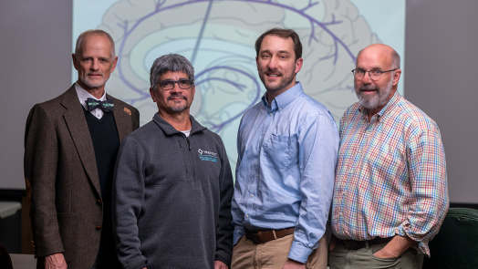 A group of four male neuroscientists stand in front of a backdrop with a picture of an animated brain on it and look at the camera.