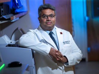A male scientist stands in a lab surrounded by computers and other lab equipment.