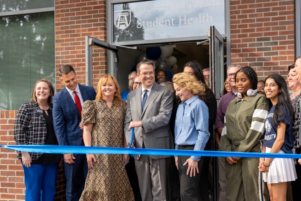 A group of men and women stand behind a ribbon with the center man holding large scissors.