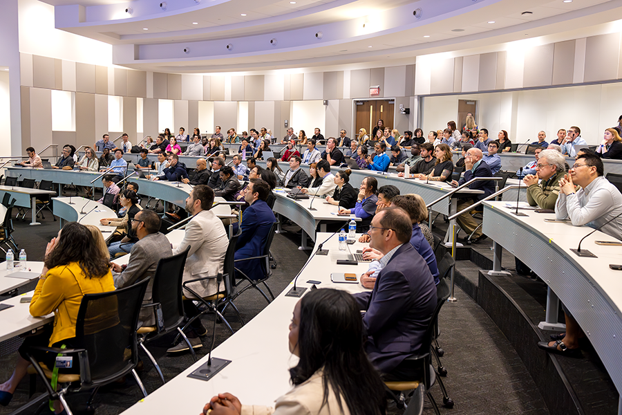 A presenter speaks to a crowded lecture hall on a modern college campus.