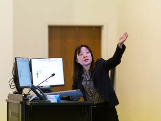 A woman presents to a large crowd in a modern lecture hall on a college campus.