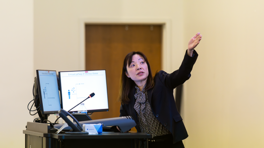 A woman presents to a large crowd in a modern lecture hall on a college campus.