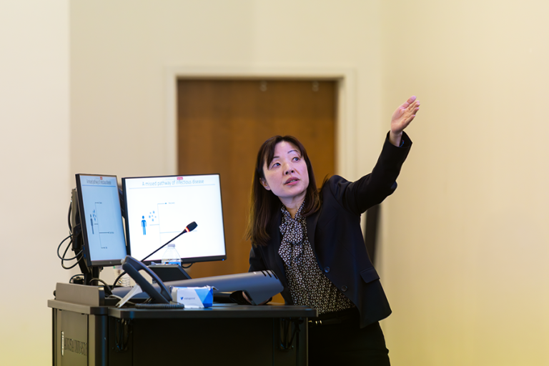 A woman presents to a large crowd in a modern lecture hall on a college campus.