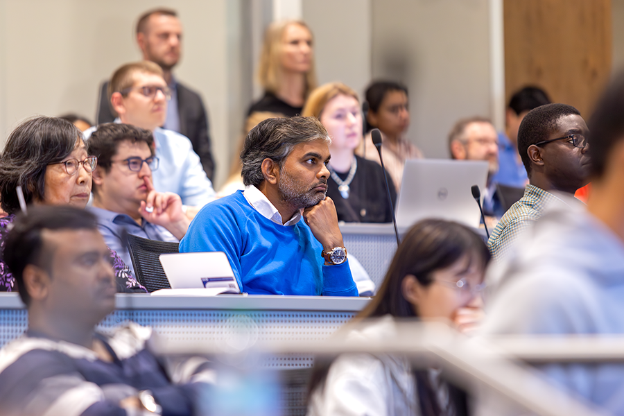 A man listens as a presenter speaks to a crowded lecture hall on a modern college campus.