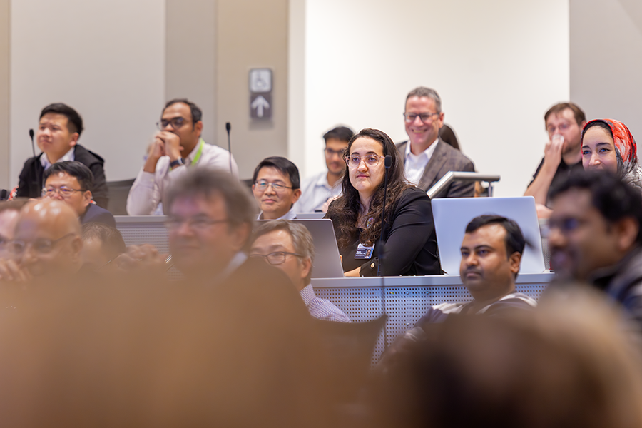 A woman listens as a presenter speaks to a crowded lecture hall on a modern college campus.