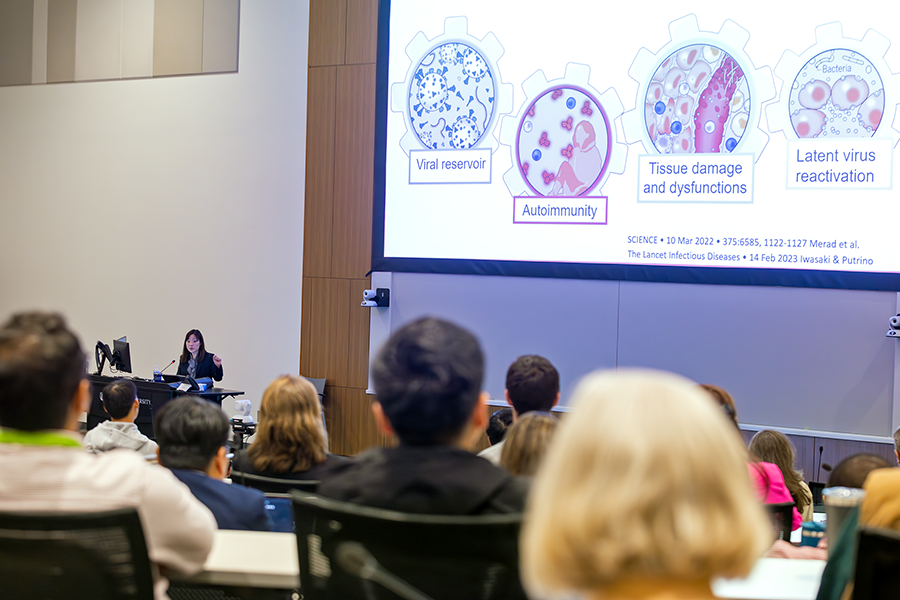 A presenter speaks to a crowded lecture hall on a modern college campus.