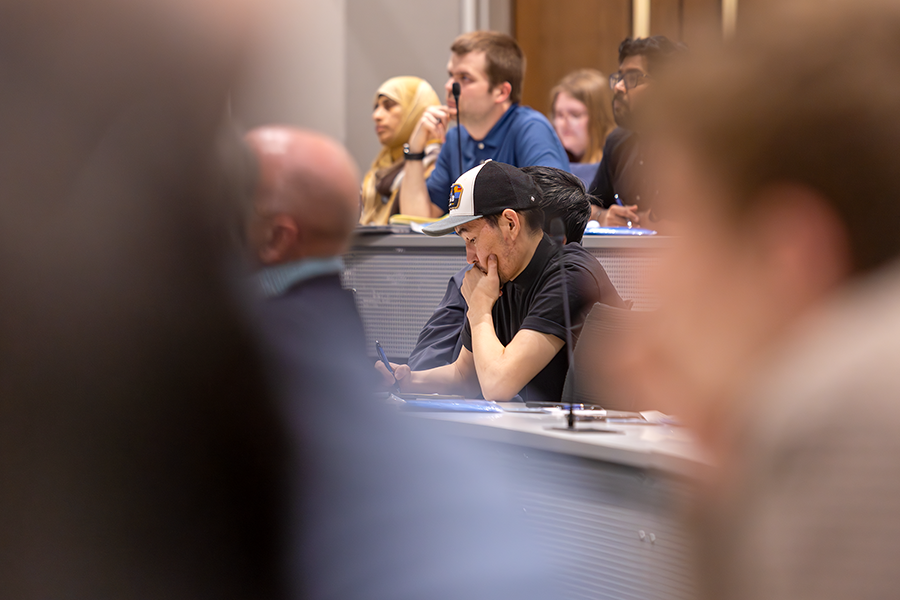 A man listens and takes notes as a presenter speaks to a crowded lecture hall on a modern college campus.