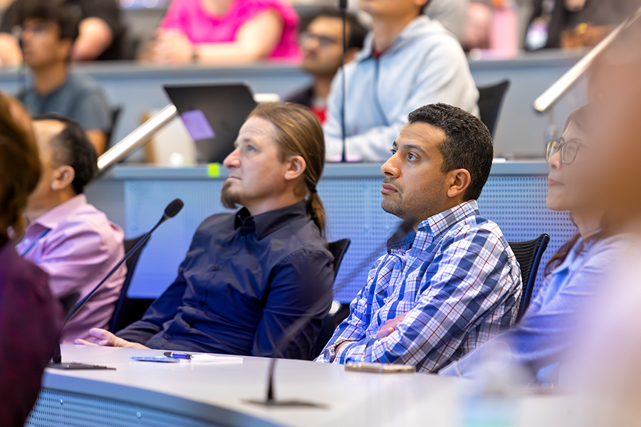 Two men listen as a presenter speaks to a crowded lecture hall on a modern college campus.