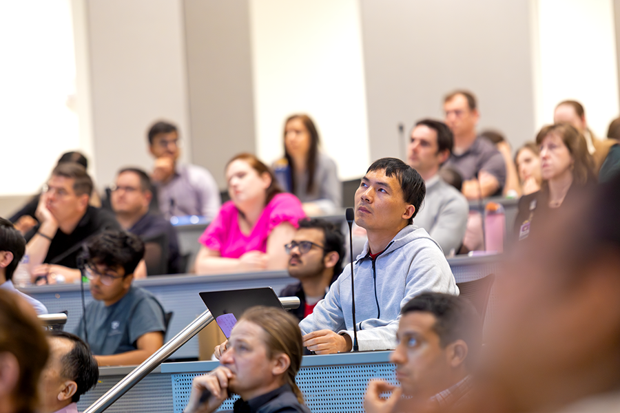 A man listens as a presenter speaks to a crowded lecture hall on a modern college campus.