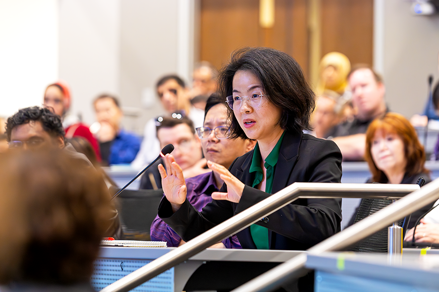A woman asks a question of a presenter during a Q&A session following a lecture in a crowded lecture hall on a modern college campus.