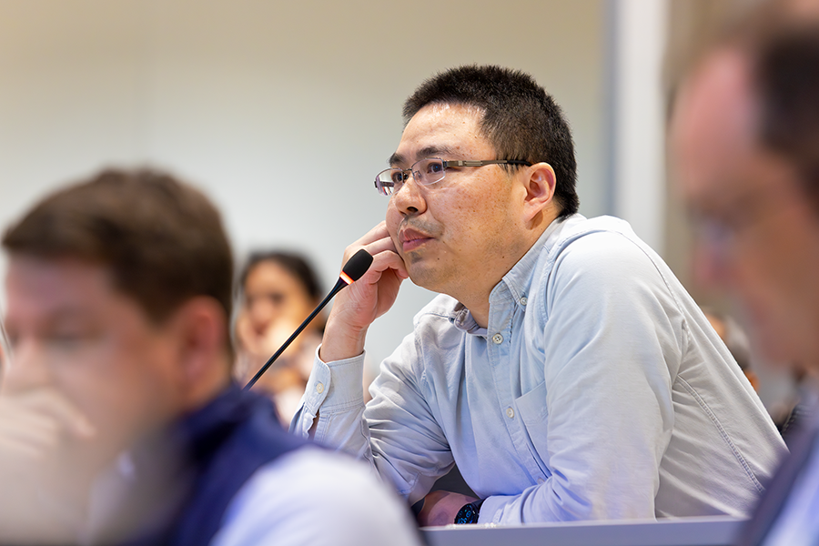 A man listens as a presenter speaks to a crowded lecture hall on a modern college campus.