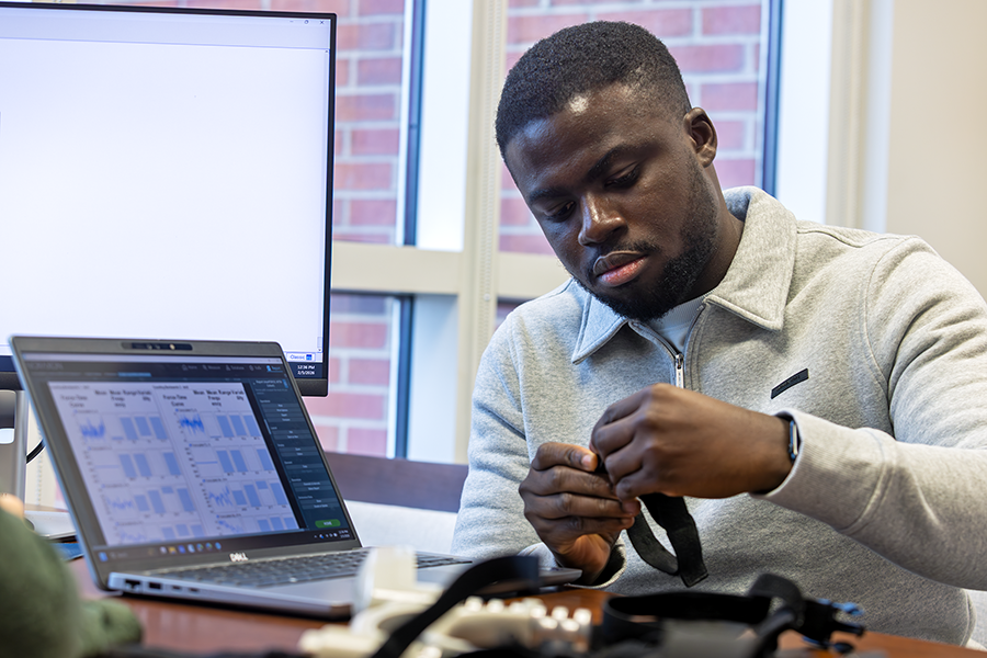 A male college student holds a wearable device and works to transfer data from the device to a laptop computer on the desk in front of him.