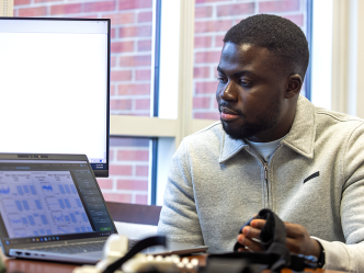 A male college student holds a wearable device and works to transfer data from the device to a laptop computer on the desk in front of him.