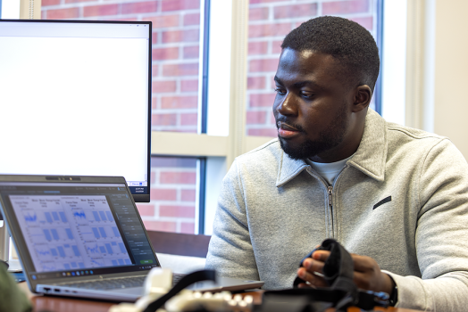 A male college student holds a wearable device and works to transfer data from the device to a laptop computer on the desk in front of him.