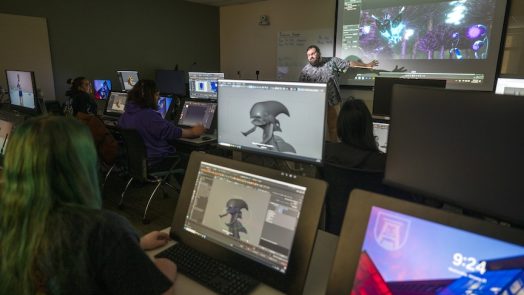 students on monitors looking at a male teacher present work on a board
