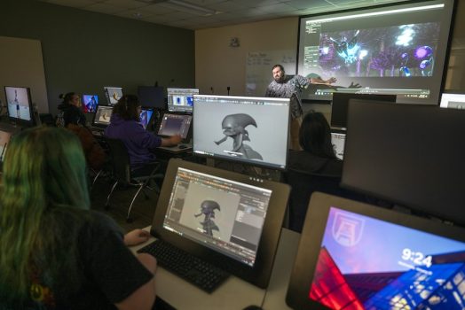 students on monitors looking at a male teacher present work on a board