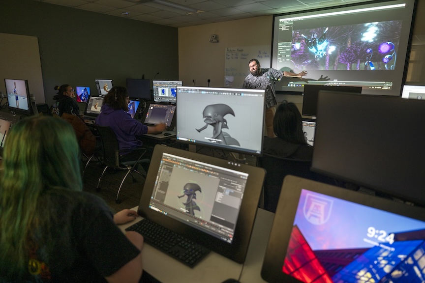 students on monitors looking at a male teacher present work on a board