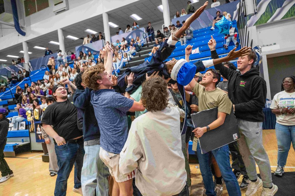 A group of men carry a man wearing a crown inside a gymnasium.