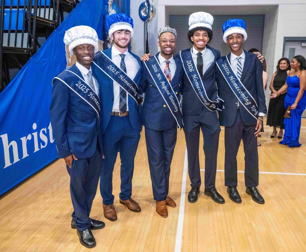 A group of men wearing crowns and sashes stand inside a gymnasium.
