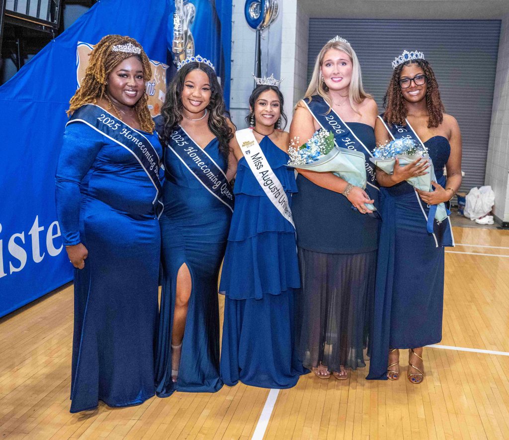 A group of men wearing crowns and sashes stand inside a gymnasium.