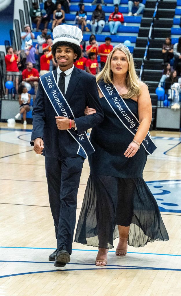 A man and woman, wearing crowns and sashes, walk arm in arm inside a gymnasium.
