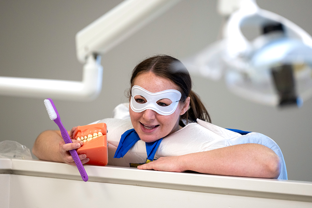 A dental hygiene student dressed in a tooth costume holds a model of a mouth and a toothbrush.