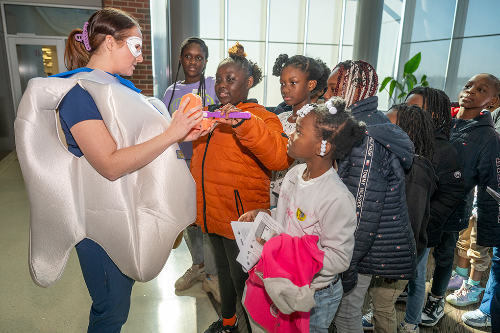 A dental hygiene student wearing a tooth costume holds a mouth model for a group of young kids to practice brushing teeth on.