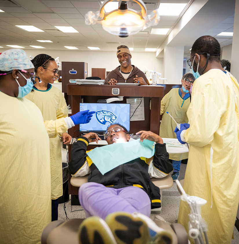 A group of dental hygiene students and a former NFL player stand around a little boy in a dental exam chair.