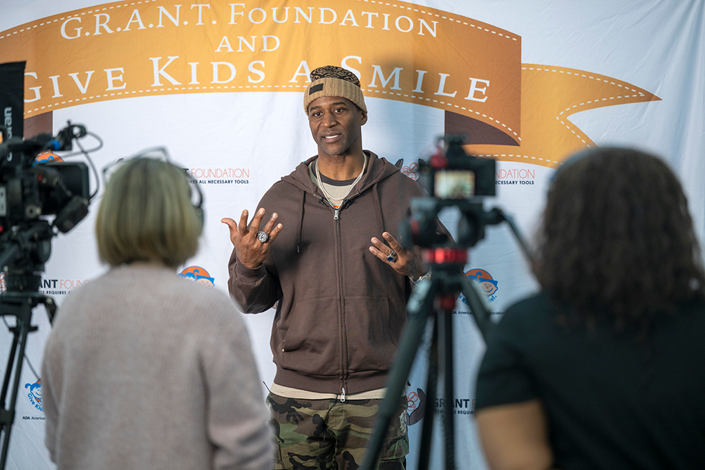 A former NFL football player stands in front of a backdrop that says "G.R.A.N.T. Foundation and Give Kids a Smile" and answers questions from journalists.