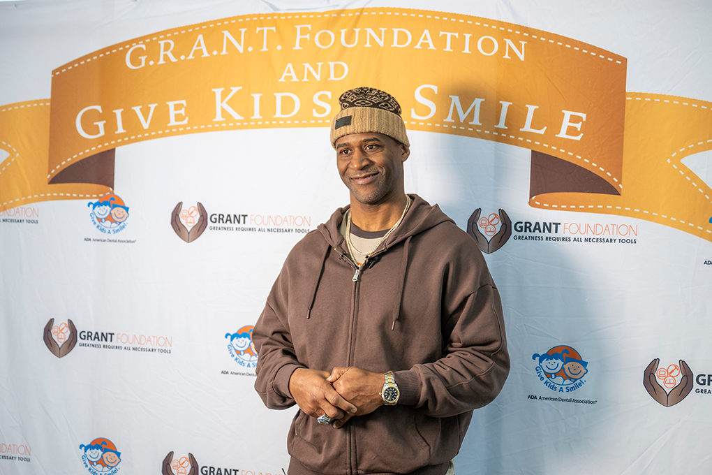 A former NFL football player stands in front of a backdrop that says "G.R.A.N.T. Foundation and Give Kids a Smile."