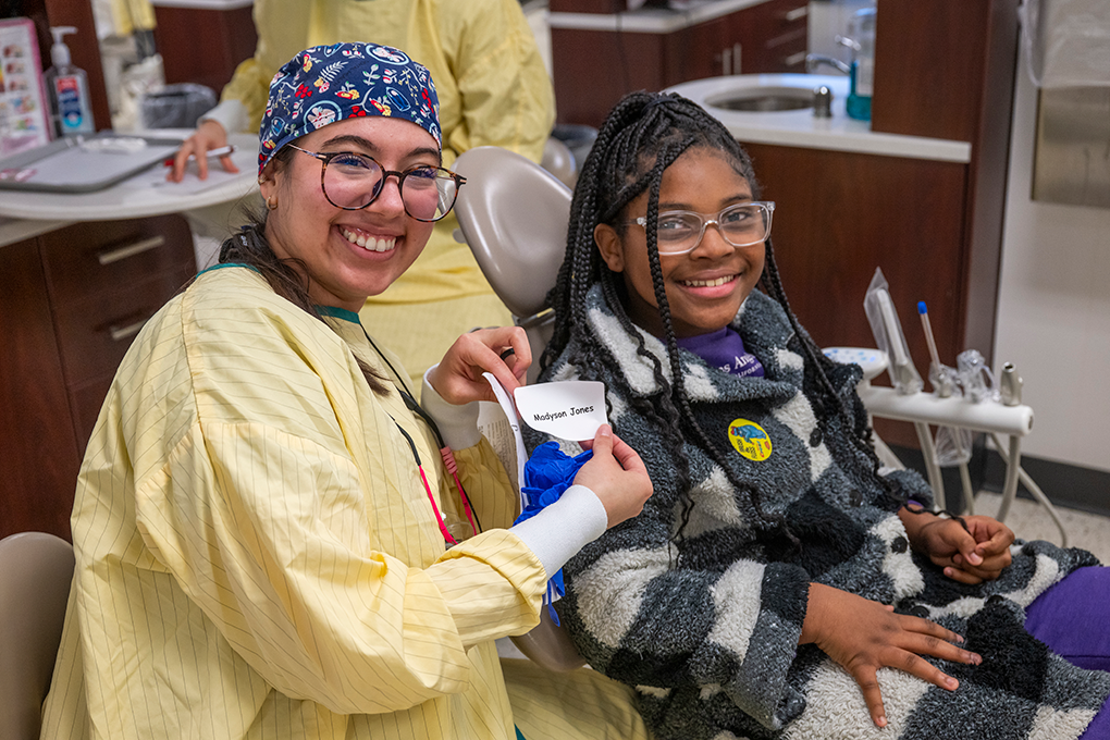 A dental hygiene student smiles for a picture with a little girl in a dental exam chair.