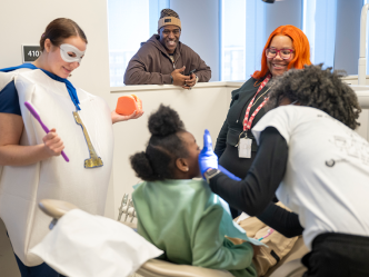 A little girl sits in a dental exam chair while a dental hygienist cleans her teeth. A teacher, a dental student wearing a tooth custom, and a former NFL football player stand by and encourage her.