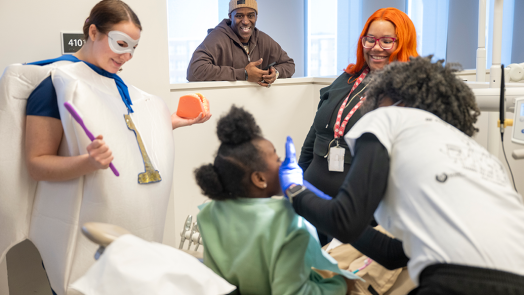 A little girl sits in a dental exam chair while a dental hygienist cleans her teeth. A teacher, a dental student wearing a tooth custom, and a former NFL football player stand by and encourage her.