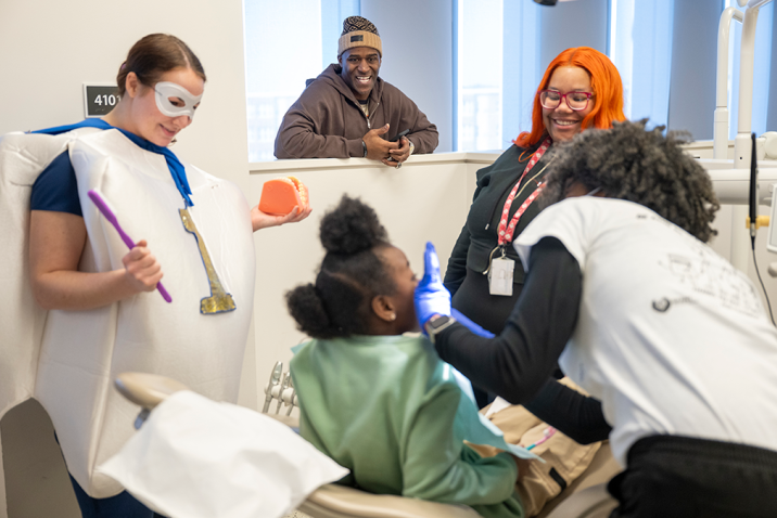 A little girl sits in a dental exam chair while a dental hygienist cleans her teeth. A teacher, a dental student wearing a tooth custom, and a former NFL football player stand by and encourage her.