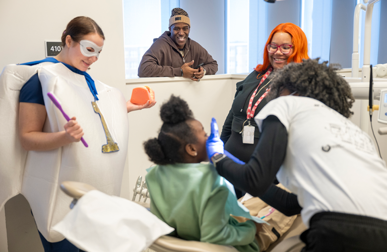 A little girl sits in a dental exam chair while a dental hygienist cleans her teeth. A teacher, a dental student wearing a tooth custom, and a former NFL football player stand by and encourage her.