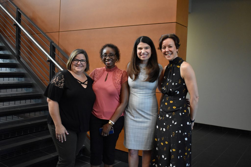 Four women in business casual attire pose together by a staircase, smiling at the camera
