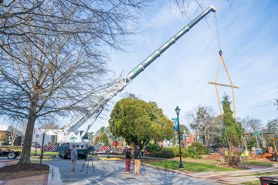 A crane lifts a large tree up in the air on the campus of a university. The crane is parked next to a fountain.