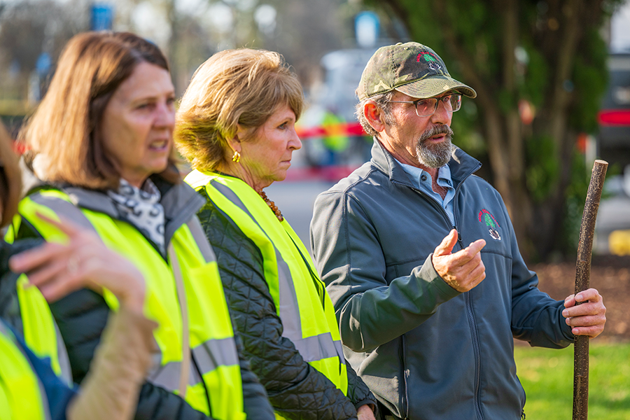 A man speaks with two women. The man is the owner of a company that is planting a fully grown tree on the campus of a university and the women are members of a local garden club. They are watching as a tree is planted.