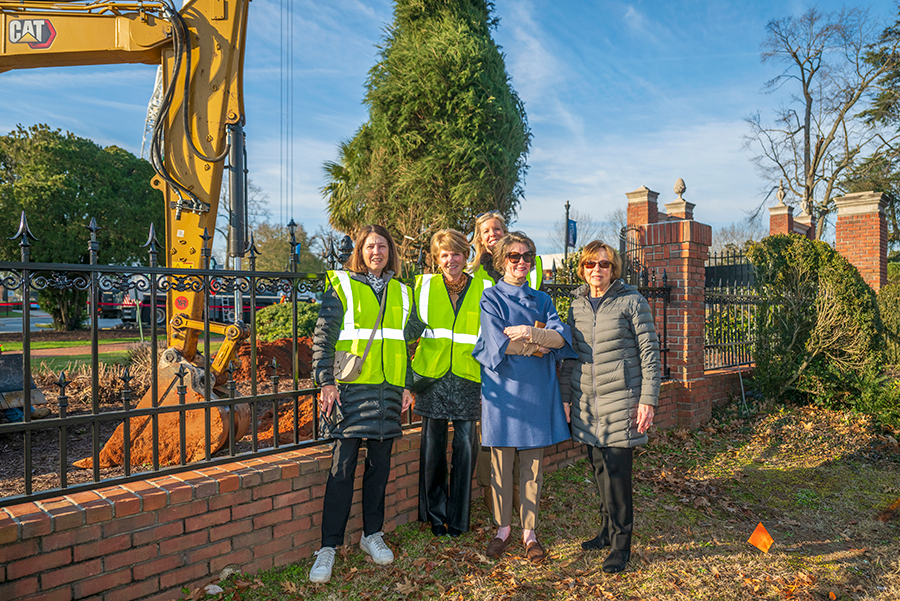 Five women stand in front of an excavator as it digs a hole to plant a full-grown tree on the campus of a university. Two of the women are wearing safety vests.