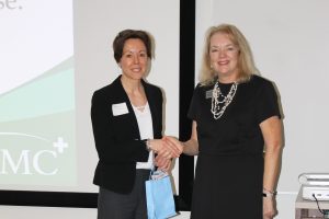 A woman in business attire shakes the hand of another woman. They are in front of a screen.