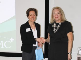 A woman in business attire shakes the hand of another woman. They are in front of a screen.