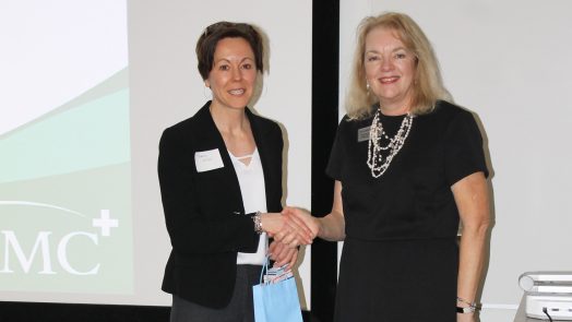 A woman in business attire shakes the hand of another woman. They are in front of a screen.