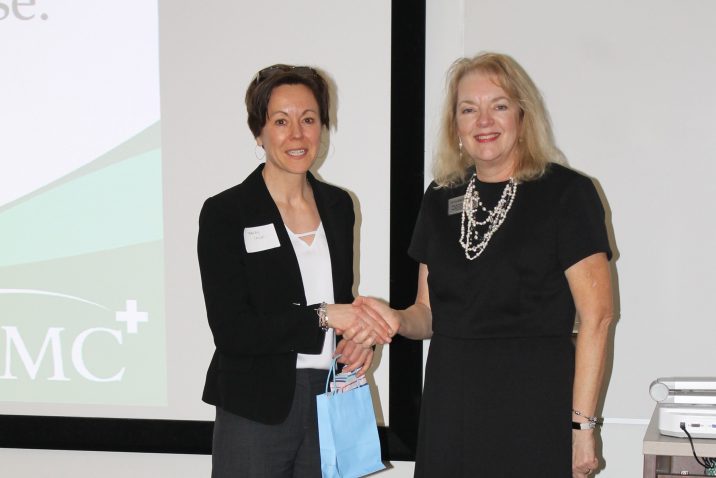 A woman in business attire shakes the hand of another woman. They are in front of a screen.
