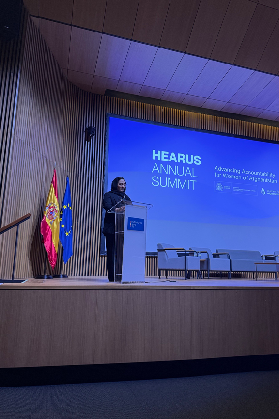 Woman presenting on stage with two flags and a presentation behind her.