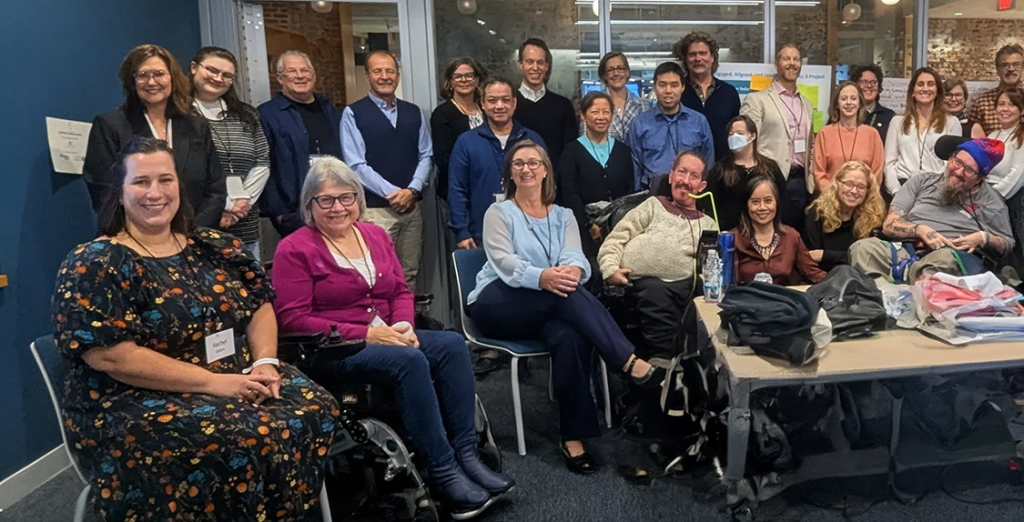A large group of people, some sitting and some standing, gather in a conference room for a meeting. Several are in wheel chairs and the group is meeting to discuss how to help those with disabilities get the care they want and deserve.