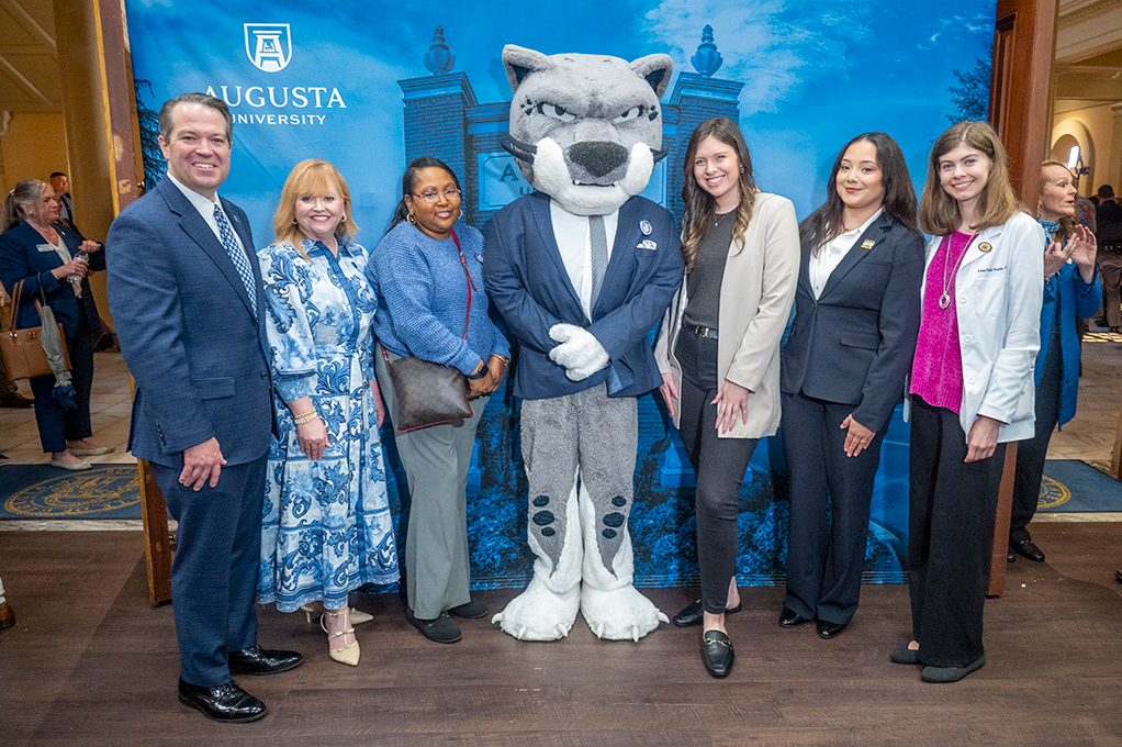 One men and five women gather in a line, smiling with a person dressed as a cat wearing a large cat head.