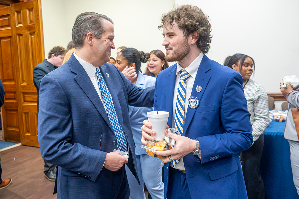 Two men smile at one another in business dress, one holding a drink and small box of fruit.