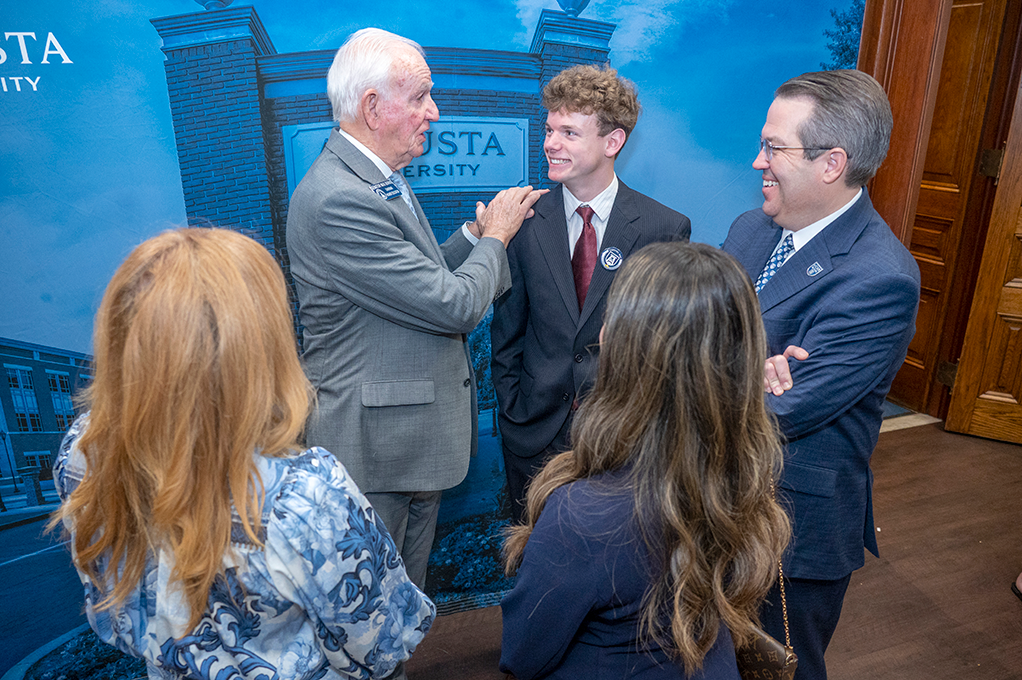 Three men and two women stand in a circle smiling with one man speaking.