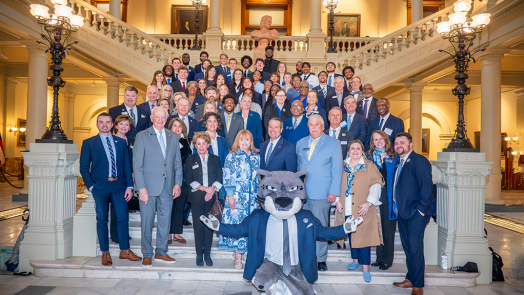 A large group of men and women various business dress pose on a very large staircase with a person wearing a cat head in the front.