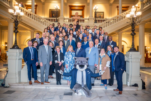 A large group of men and women various business dress pose on a very large staircase with a person wearing a cat head in the front.